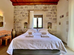 A bedroom with a stone wall, a wooden bed frame, a bedside table, and a window with curtains.