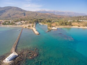 An aerial view of a coastal area with clear turquoise waters, a white building near the shore, and mountains in the background. - Villa Olivia in Vrises (Apokoronas)