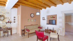 Een gezellige woonkamer met een houten plafond, een stenen muur, een bank, een rode fauteuil, een witte stoel, een televisie en een klein tafeltje met een lamp.