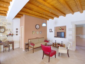 Un salon chaleureux avec un plafond en bois, un mur en pierre, un canapé, un fauteuil rouge, un fauteuil blanc, un téléviseur et une petite table avec une lampe.