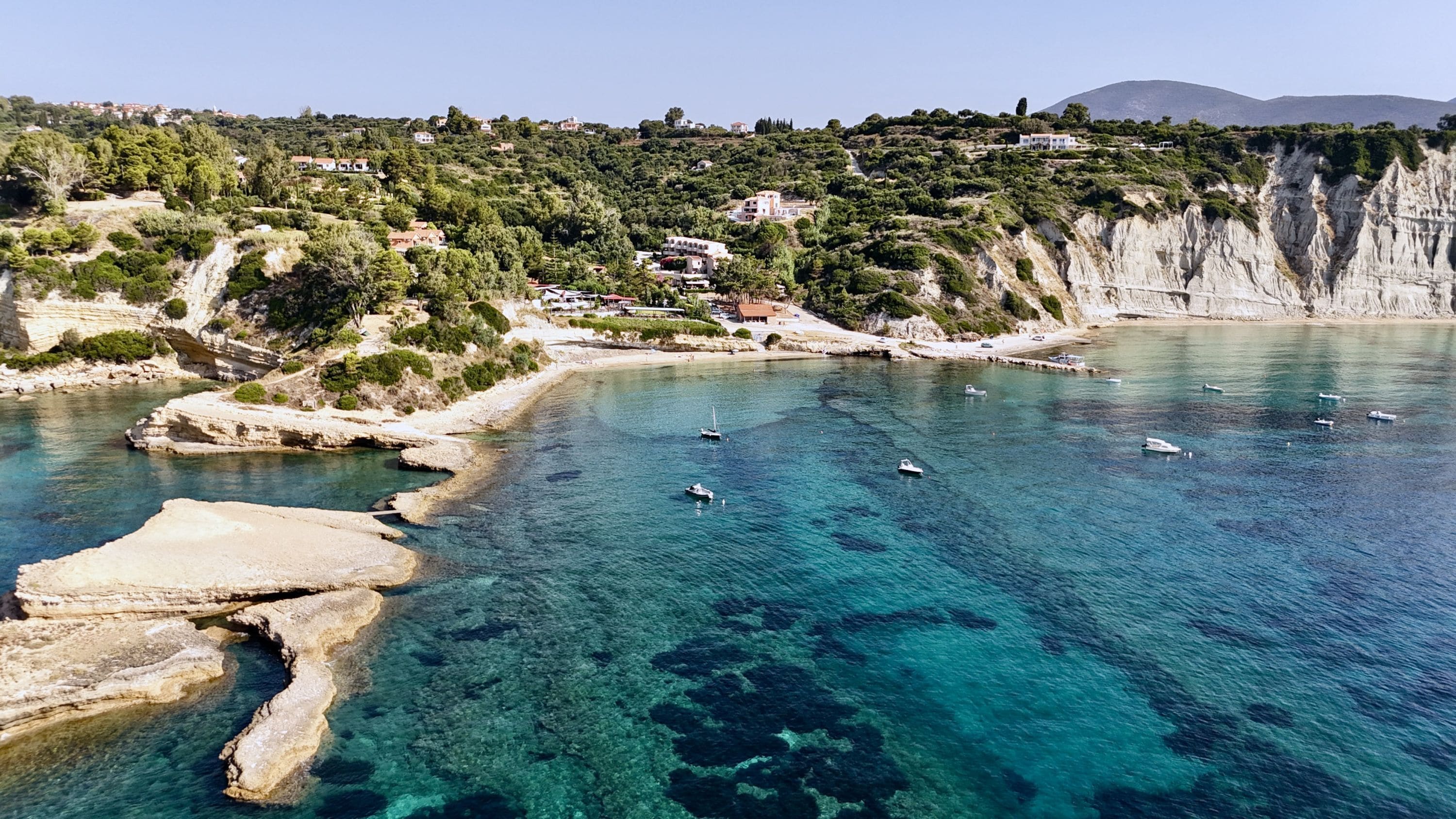 A coastal area with clear blue waters, rocky formations, and buildings on a hillside.