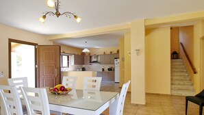 A dining area with a glass table and white chairs, a kitchen in the background, and a staircase leading to another floor. - Villa Alexandros in Spartia (Kefalonia)