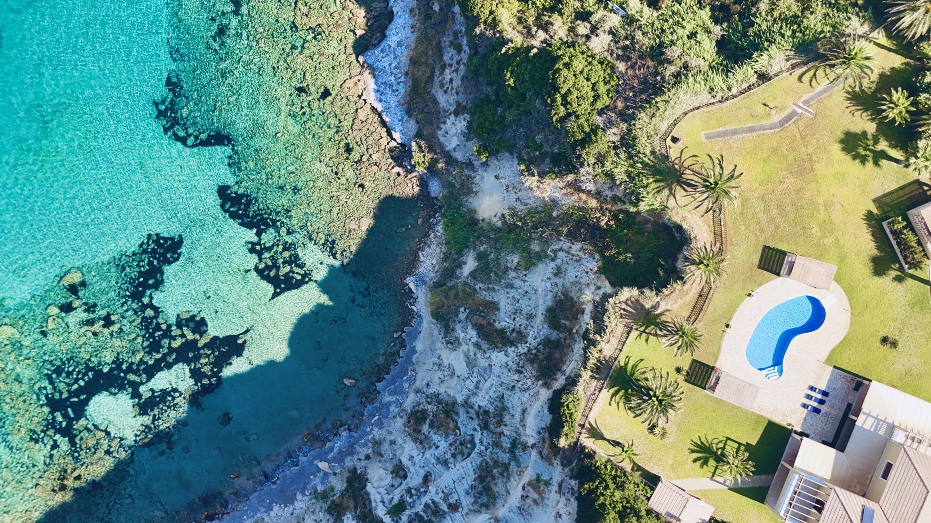 Oversigtsbillede af en ejendom ved kysten med swimmingpool, frodig vegetation og en klippefyldt kystlinje.