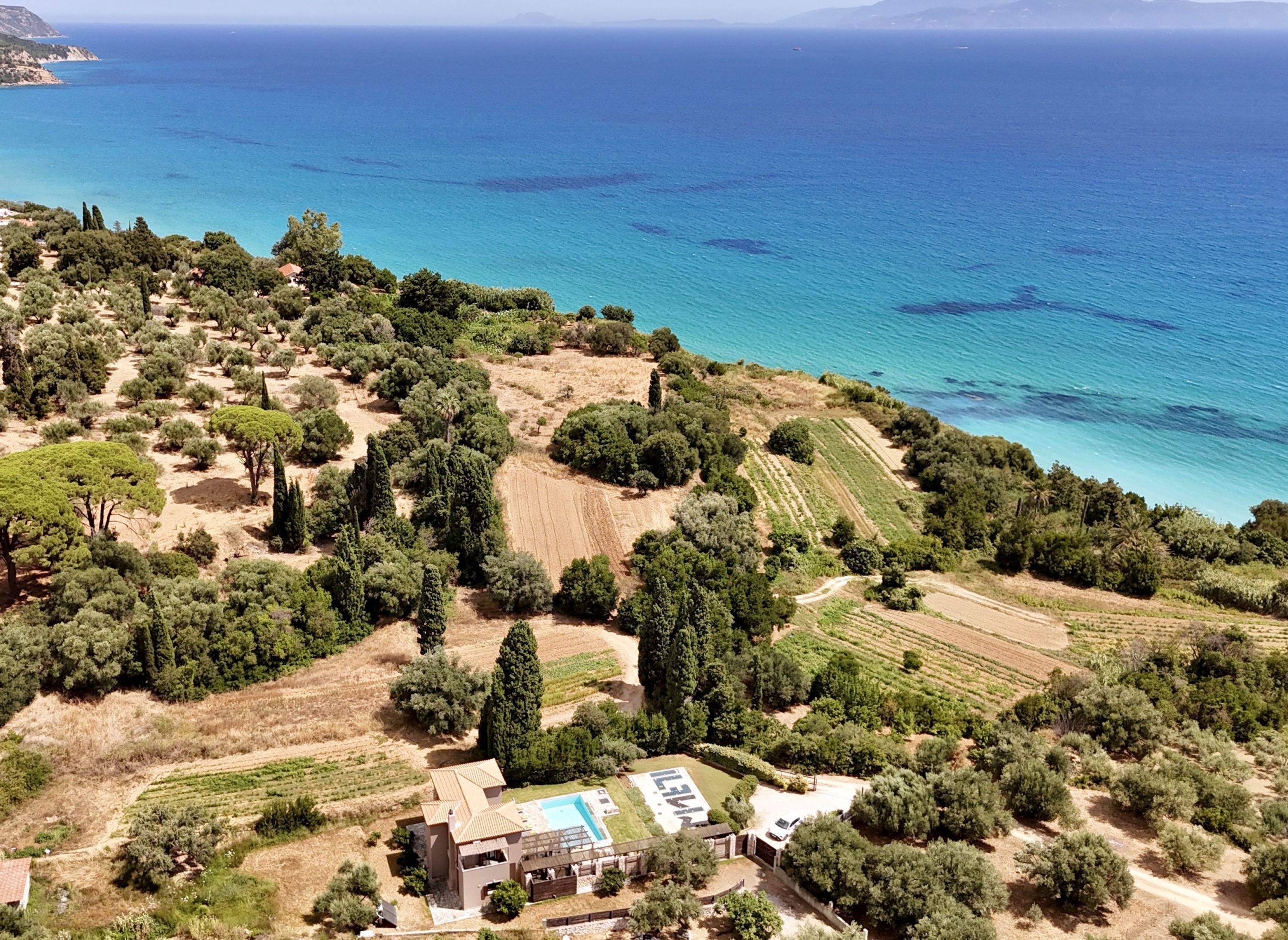 An aerial view of a property with a swimming pool, terraced fields, and a coastal backdrop.