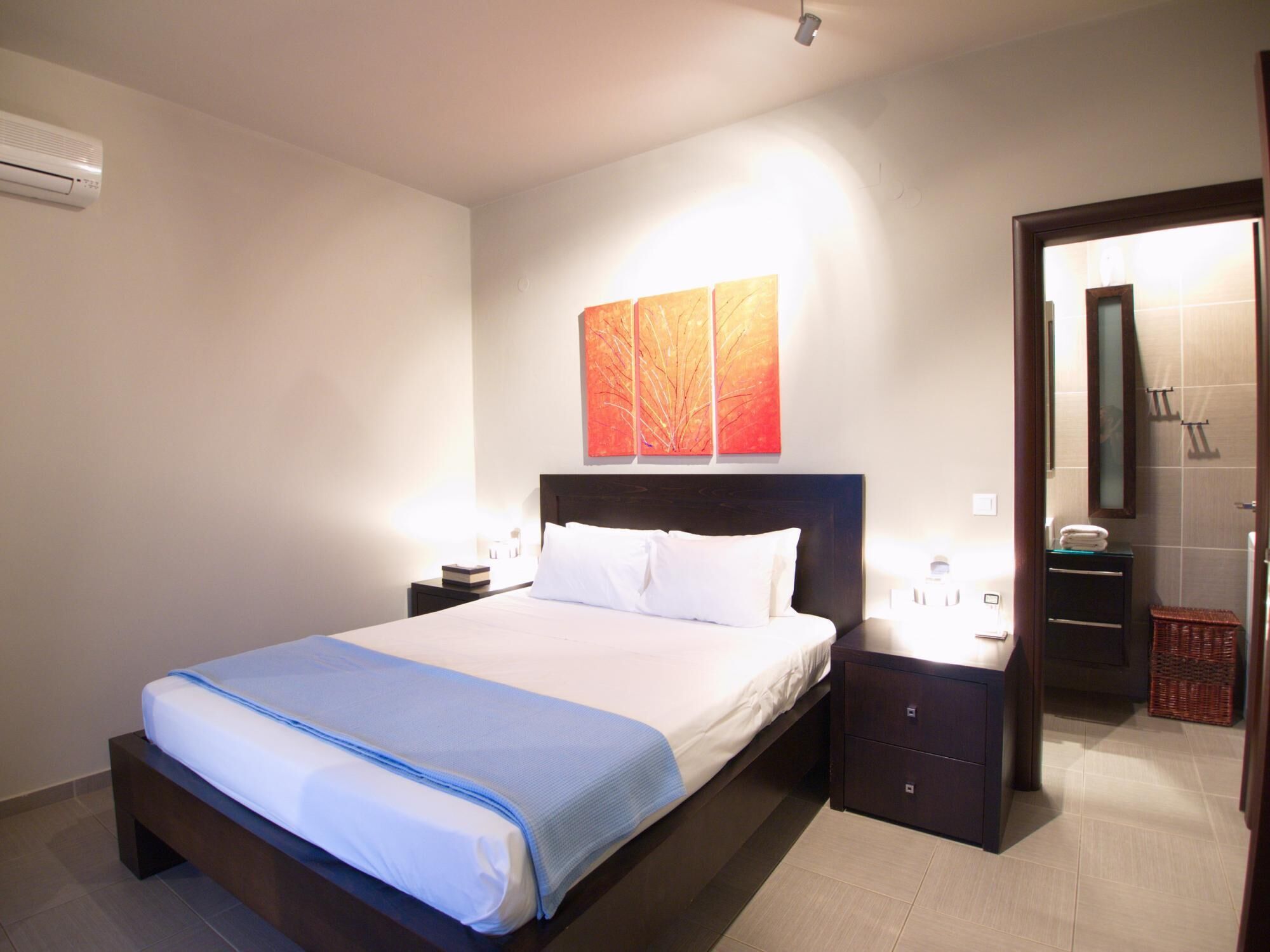 A hotel room with a bed, bedside tables, a nightstand, a wall-mounted air conditioner, a mirror, and a door leading to a bathroom.