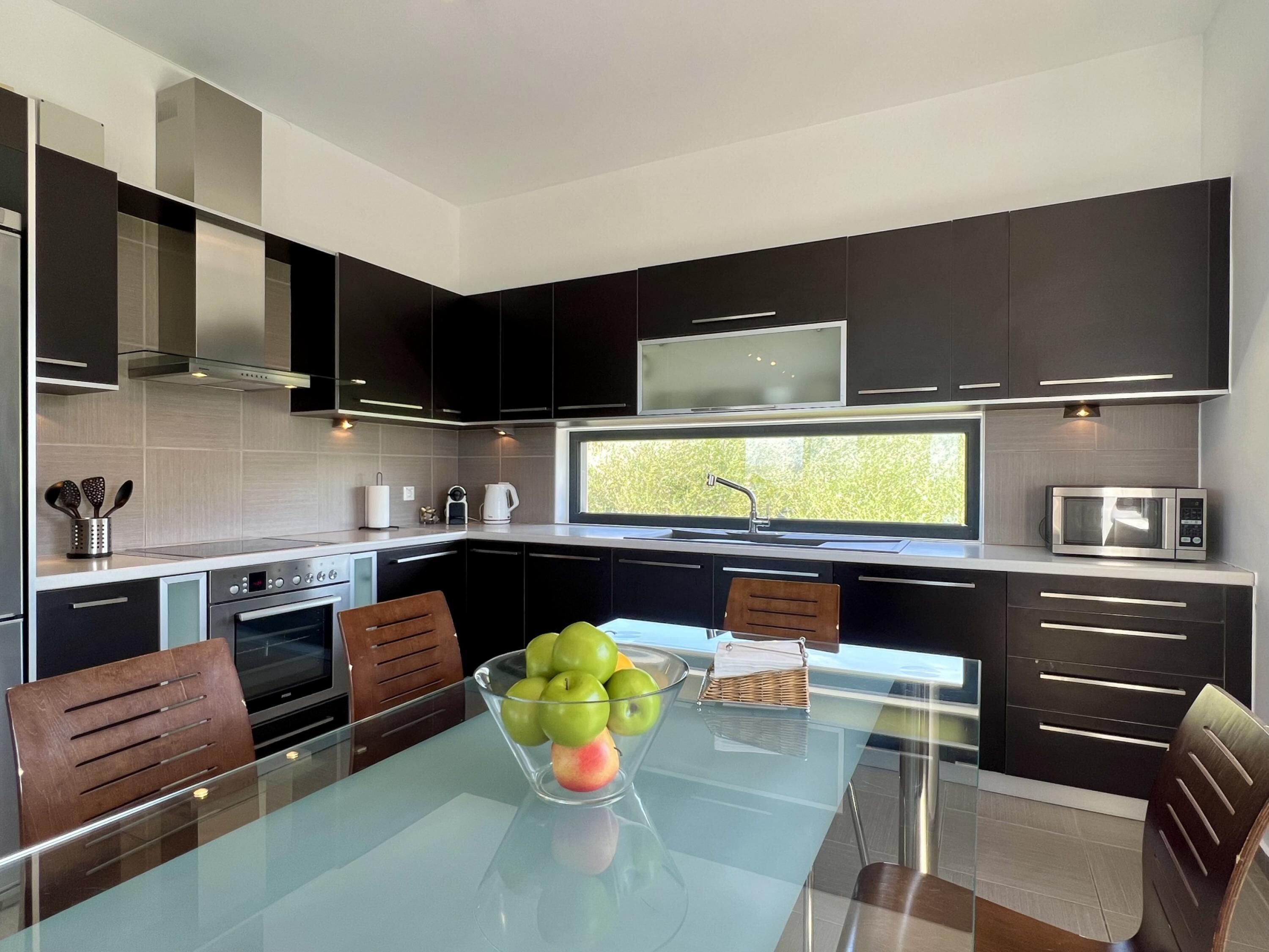 A modern kitchen with dark cabinetry, a glass-top dining table, and a bowl of fruit.