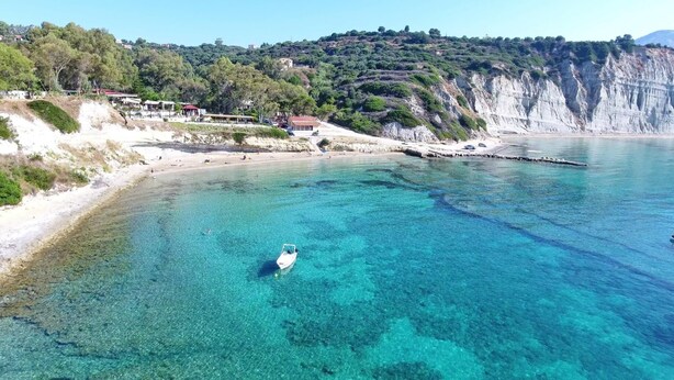 A coastal area with clear turquoise waters, a boat, and a beach with buildings in the background. - Villa Emiliata in Spartia (Kefalonia)