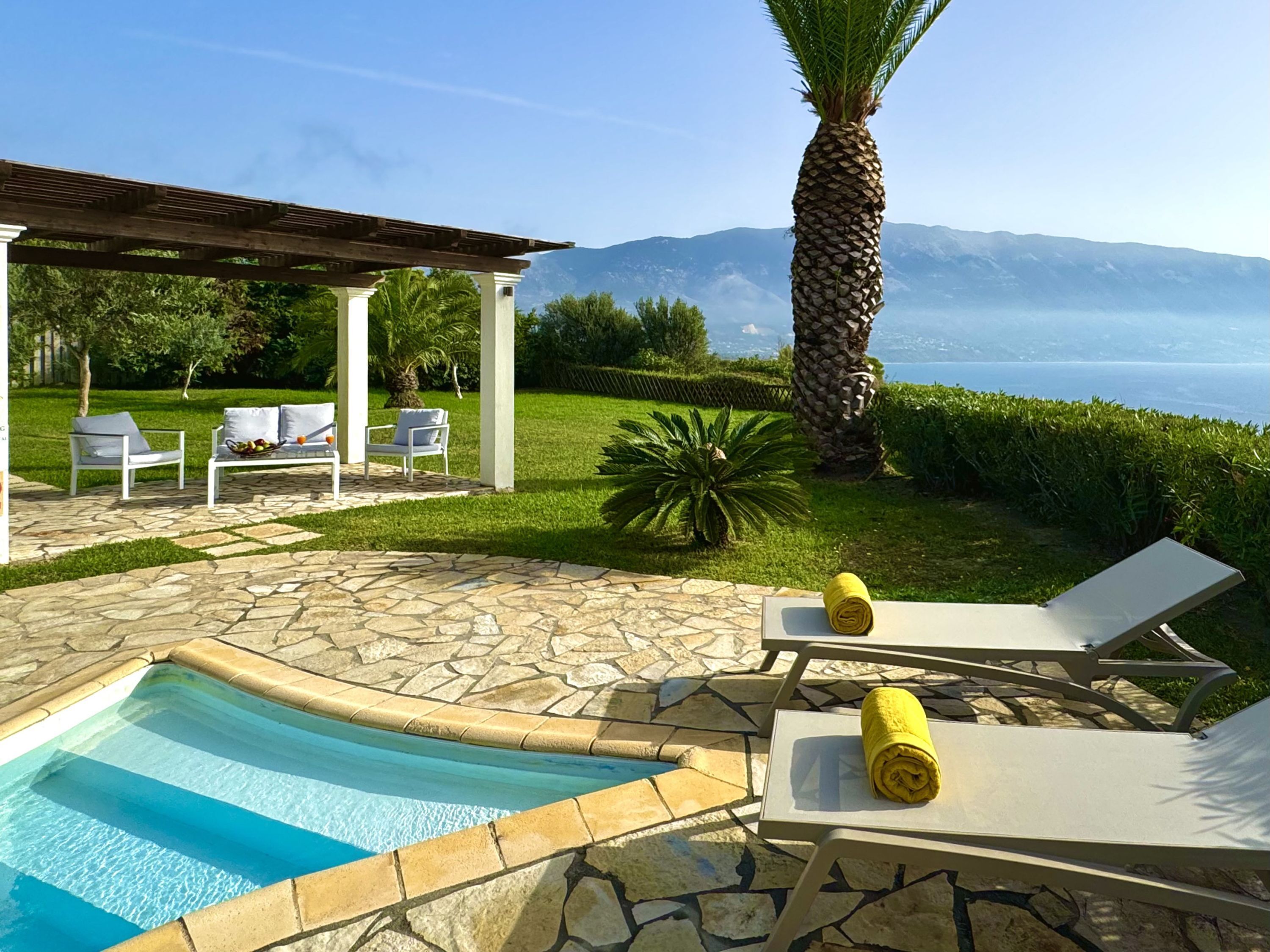 A pool area with stone tiles, lounge chairs, a pergola, and a view of the sea and mountains.