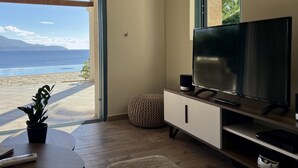 A modern living room with a large TV, wooden flooring, a round table with a plant, and a view of the ocean. - Villa Penelope in Spartia (Kefalonia)