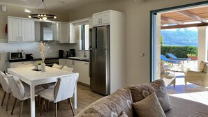 A modern kitchen with white cabinets, a stainless steel refrigerator, and a dining area with a white table and chairs. - Villa Penelope in Spartia (Kefalonia)