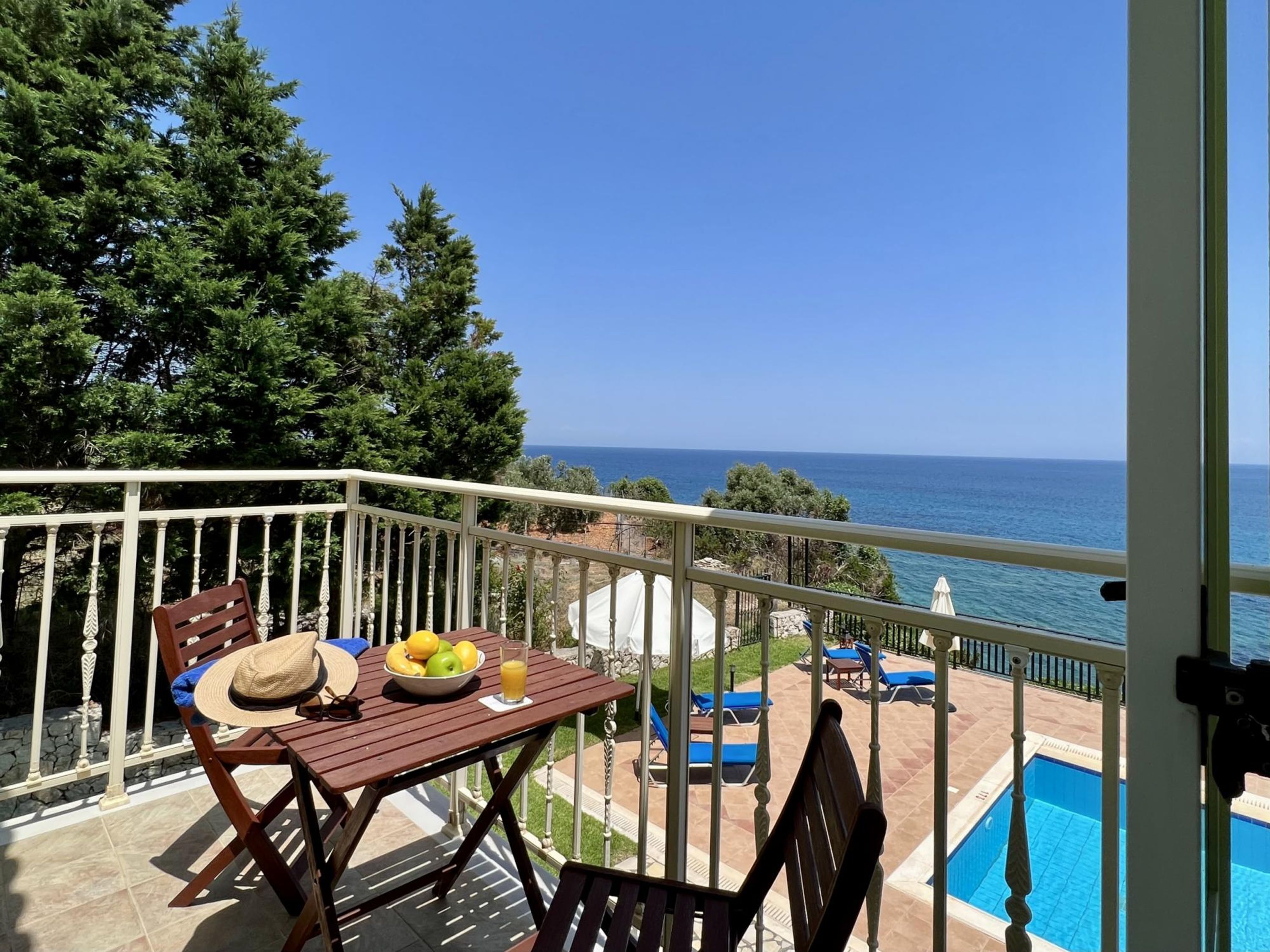 A balcony with a wooden table, a bowl of fruit, and a glass of juice. There are lounge chairs and a pool in the background.