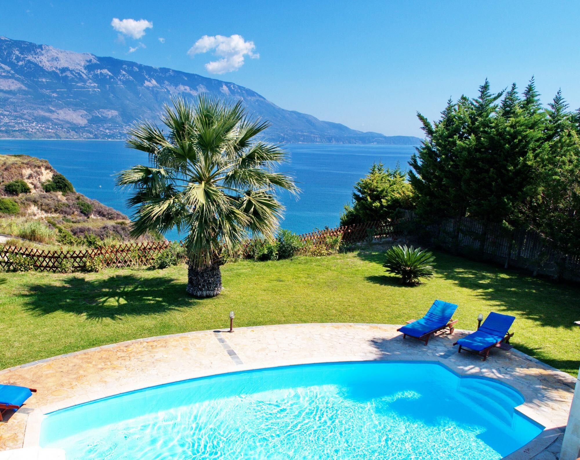 A swimming pool with two blue lounge chairs, a palm tree, and a view of the sea and mountains.