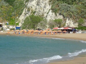 A beach with rows of sun loungers and umbrellas, a rocky cliff in the background, and a clear view of the sea. - Villa Vangelis in Stylos (Apokoronas)