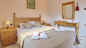 A neatly made bed with a quilted bedspread, a wooden headboard, and two bedside tables with lamps. A mirror and a vanity with a towel and a red cloth are also visible. - Maros Spitaki in Varipetro (Chania)