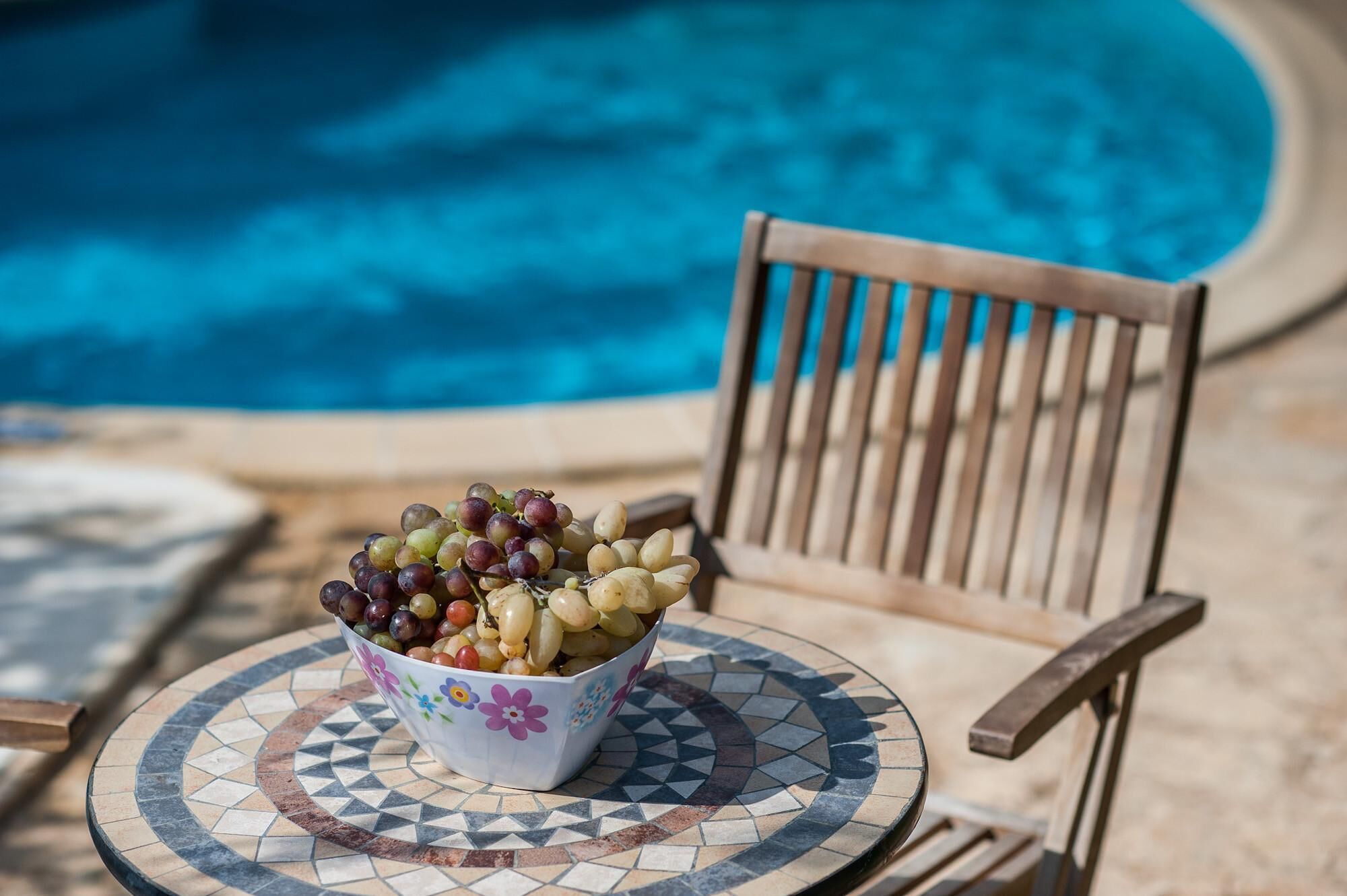 Un saladier rempli de fruits variés, posé sur une table carrelée, à côté d’une chaise au bord de la piscine.