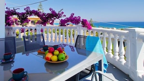 Une table avec un panier de fruits, deux tasses et une serviette bleue, sur un balcon donnant sur la mer.