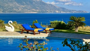 A poolside area with lounge chairs and a swan statue, overlooking a mountainous coastline. - Villa Dionysos in Spartia (Kefalonia)