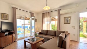 A living room with a TV, sofa, and a glass table. There is a sliding glass door leading to a balcony with a view of the ocean. - Villa Dionysos in Spartia (Kefalonia)