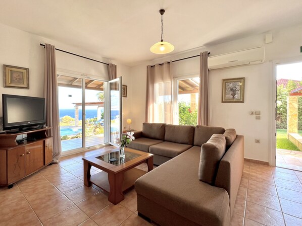 A living room with a TV, sofa, and a glass table. There is a sliding glass door leading to a balcony with a view of the ocean. - Villa Dionysos in Spartia (Kefalonia)