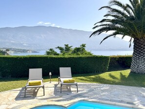 A poolside area with two lounge chairs, a palm tree, and a view of a lake and mountains. - Villa Hephaestus in Spartia (Kefalonia)