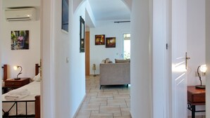A hallway leading to a living room with a sofa, a bed with a nightstand, and a lamp. There is a painting on the wall and an air conditioning unit above the bed. - Villa Hephaestus in Spartia (Kefalonia)