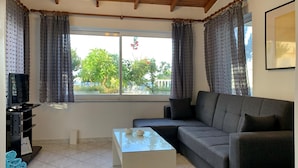 A living room with a grey sofa, a small coffee table, and a TV on a stand. There are curtains on the windows, and a view of greenery outside. - Rouladina Apt 4 in Stalos (Chania)