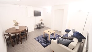 Un salon avec un canapé blanc, une petite table basse, une table à manger avec des chaises, un téléviseur fixé au mur et une plante en pot.