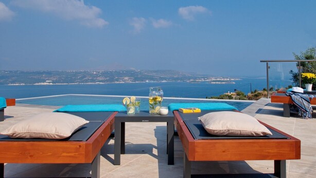 A terrace with two wooden lounge chairs, a table with a glass of water and lemon slices, and a view of the sea and distant land. - Villa Nikos in Megala Chorafia (Chania)