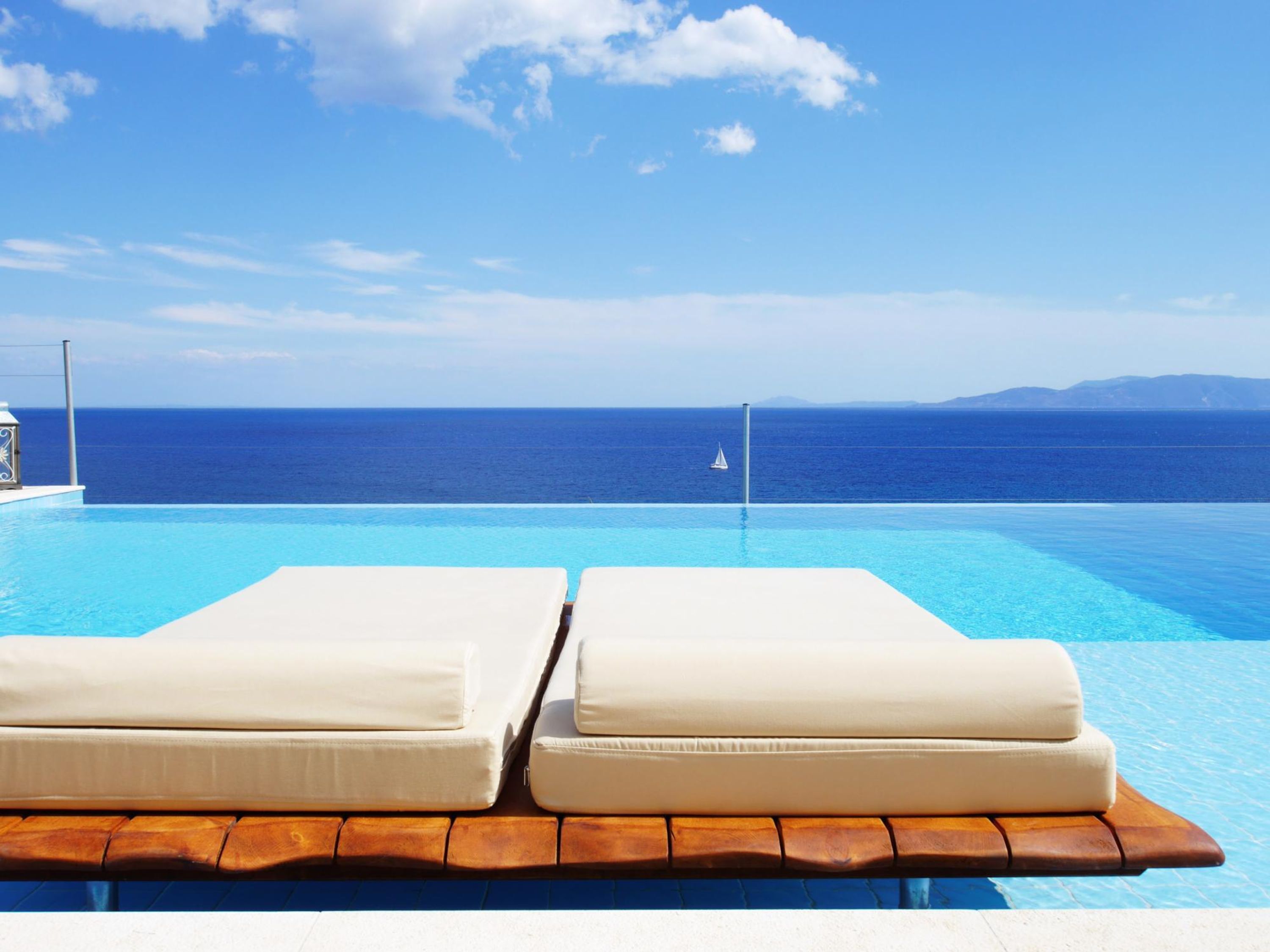 Een zwembadterras met twee witte loungestoelen die uitkijken op een heldere blauwe zee onder een zonnige lucht.