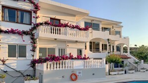 Een meerlaags hotel met een zwembad, balkons met witte relingen en roze bloemen.
