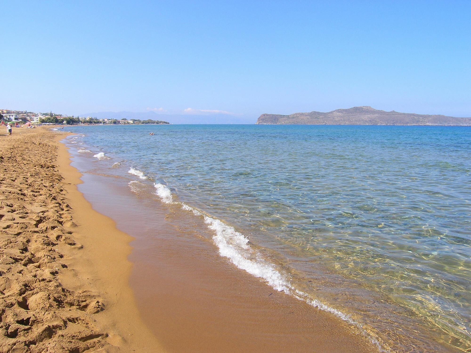 Ein Strand mit klarem Wasser, sandigem Ufer und Bergen in der Ferne.