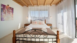 A bedroom with a wooden ceiling, a large bed with a metal frame, two bedside tables, and a window with sheer curtains. - Villa Mounda Apollon (Kefalonia)