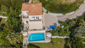 An aerial view of a two-story house with a swimming pool, surrounded by lush greenery and a paved driveway. - Villa Mounda Apollon (Kefalonia)
