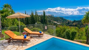 A poolside area with two lounge chairs and a table, overlooking a scenic landscape with trees and a view of the sea. - Villa Mounda Apollon (Kefalonia)