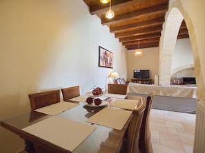 Uma sala de jantar com uma mesa de madeira, cadeiras e uma tigela de frutas. Há uma televisão e uma lareira ao fundo.