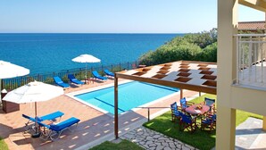 A pool area with blue lounge chairs and umbrellas, a checkered sunshade, and a view of the sea. - Villa Sakos in Skala (Kefalonia)