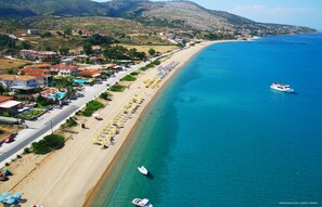 A coastal town with a long sandy beach lined with umbrellas and sun loungers, a marina with boats, and buildings with red roofs.