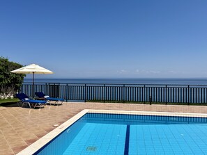 A swimming pool with a view of the ocean, a railing, and two lounge chairs with an umbrella. - Villa Sakos in Skala (Kefalonia)