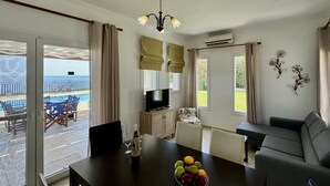 A living room with a dining table, a sofa, a TV, and a view of the sea. - Villa Sakos in Skala (Kefalonia)