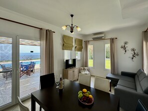 A living room with a dining table, a sofa, a TV, and a view of the sea. - Villa Sakos in Skala (Kefalonia)