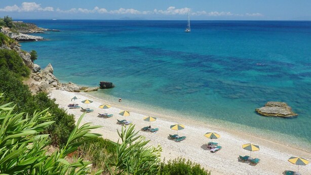 A beach with clear turquoise water, a sailboat in the distance, and several beach umbrellas. - Villa Sakos in Skala (Kefalonia)