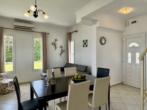 A dining area with a table, chairs, and a chandelier. There is a clock on the wall and a door with a handle. - Villa Sakos in Skala (Kefalonia)