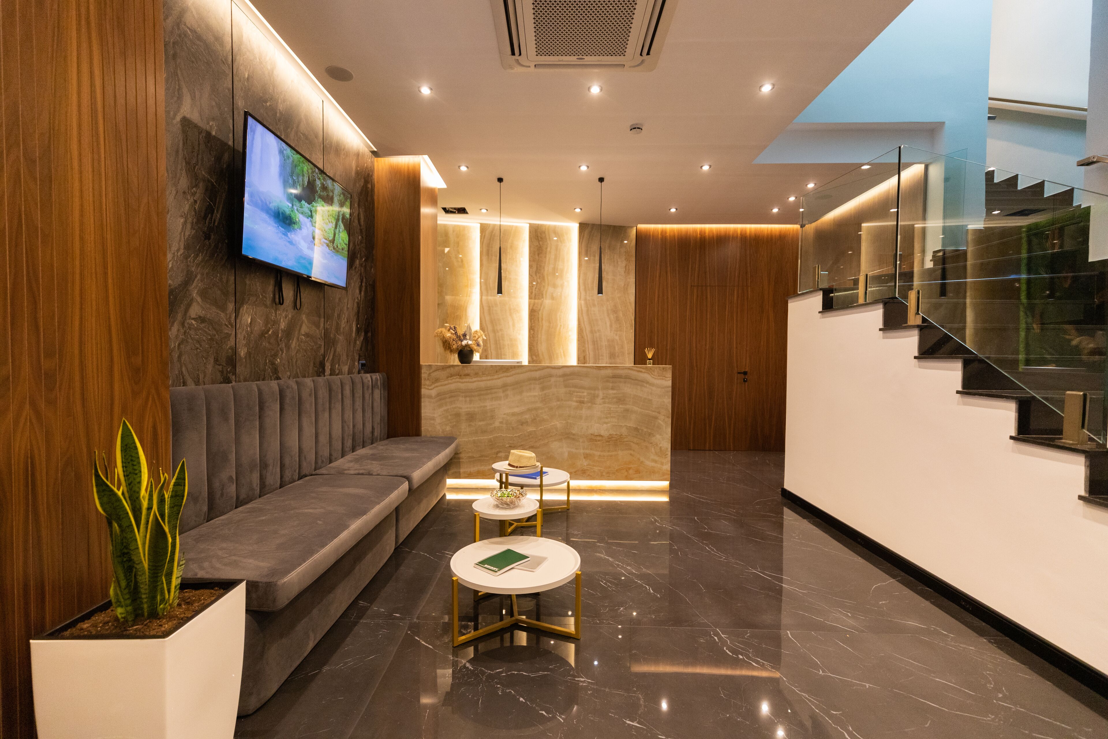 Un hall d’hôtel moderne avec un comptoir d’accueil, un canapé, deux tables rondes et un grand téléviseur à écran plat fixé au mur.