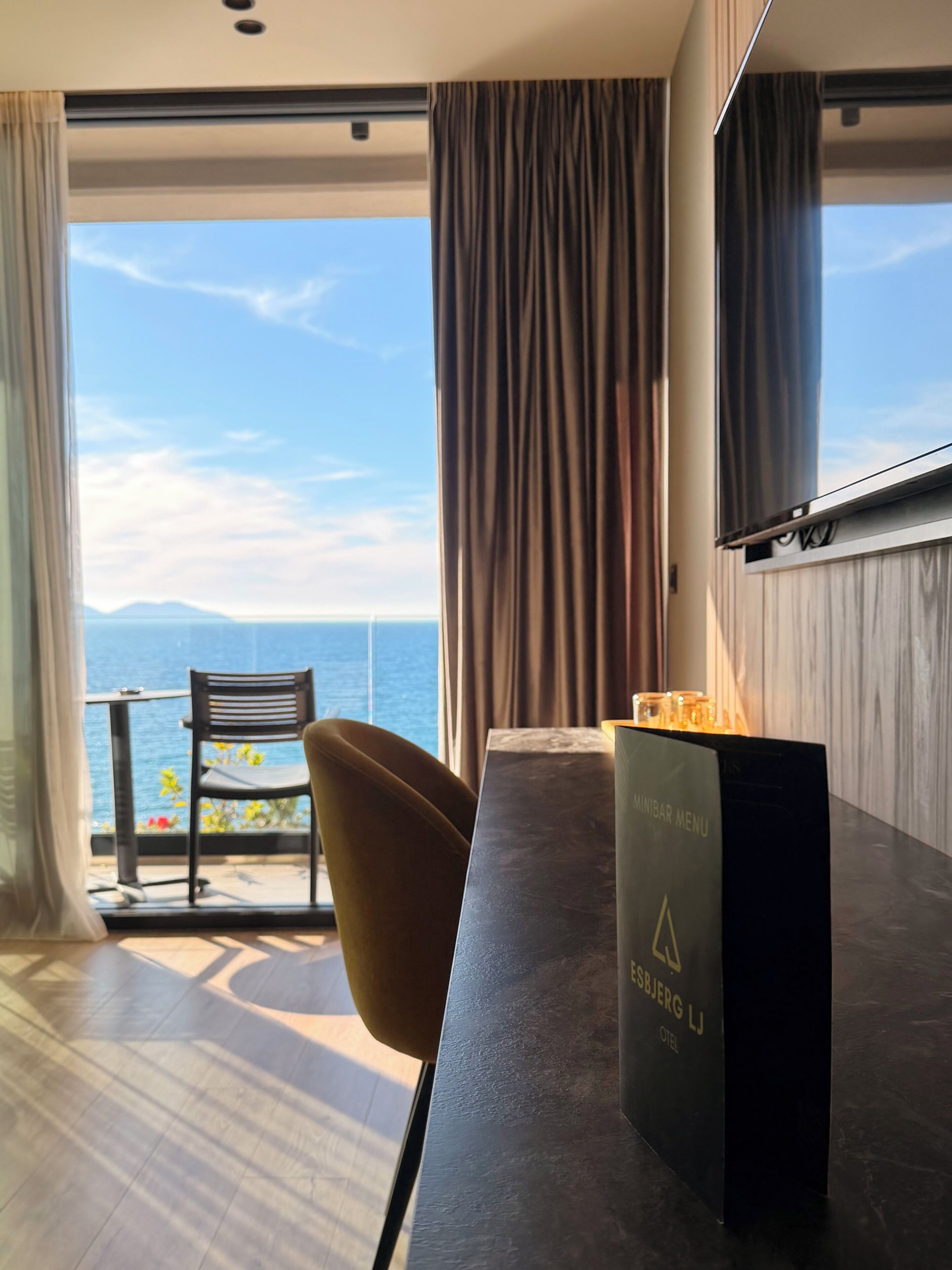A modern hotel room with a large window, a dining table, a chair, and a minibar.