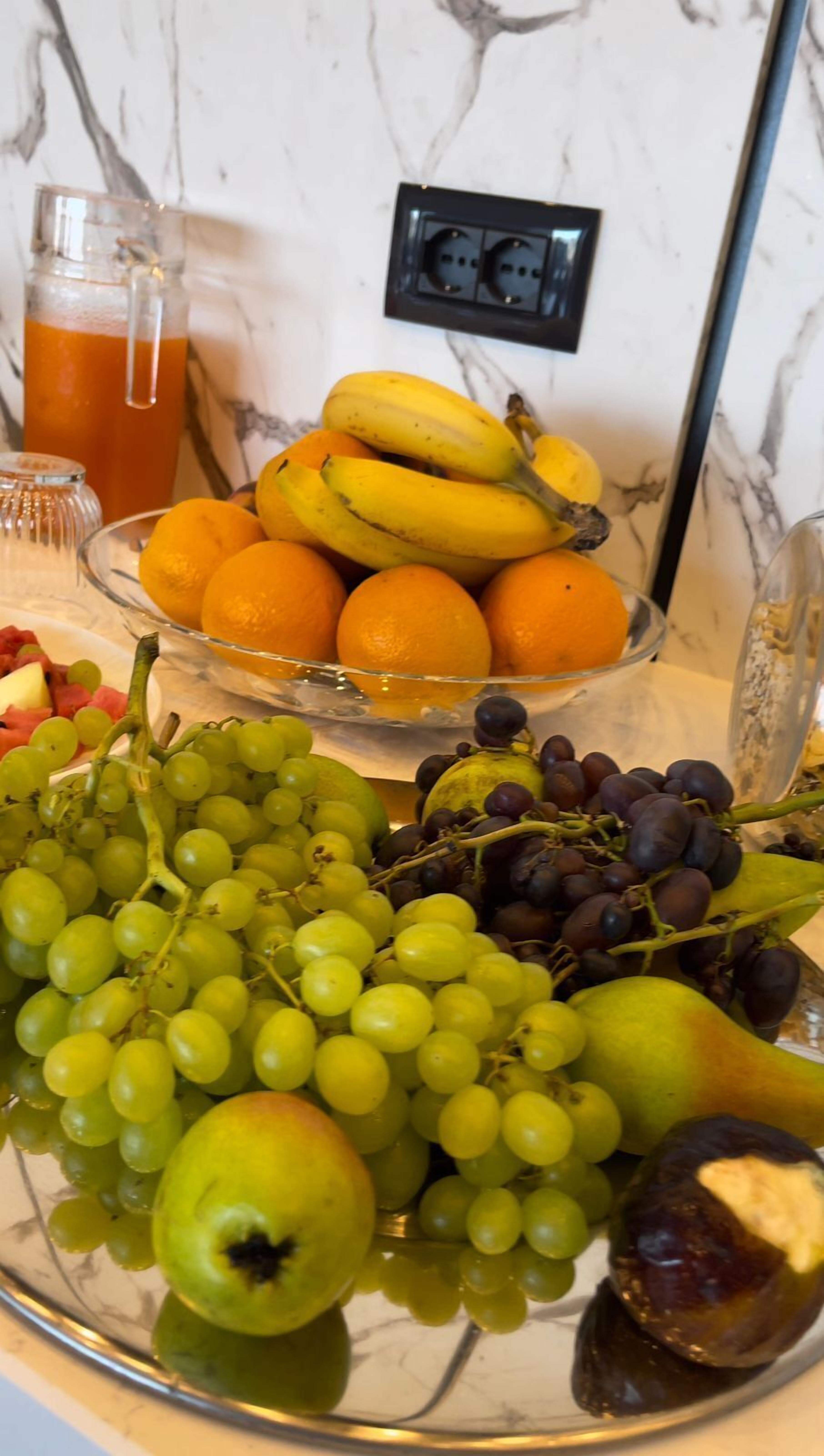 Una bandeja de frutas con plátanos, naranjas, uvas y una pera sobre una encimera de mármol.