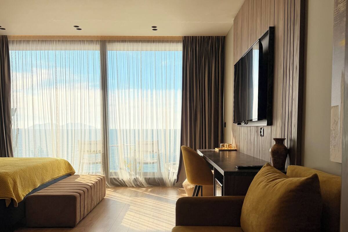 Une chambre d’hôtel moderne dotée d’une grande fenêtre, d’un téléviseur à écran plat fixé au mur, d’un canapé, d’un bureau et d’un lit recouvert d’une couverture jaune.