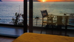 A room with a large window overlooking a sunset over the sea, a table with chairs, and a bed with a yellow blanket. - Esbjerg Hotel (Vlorë)