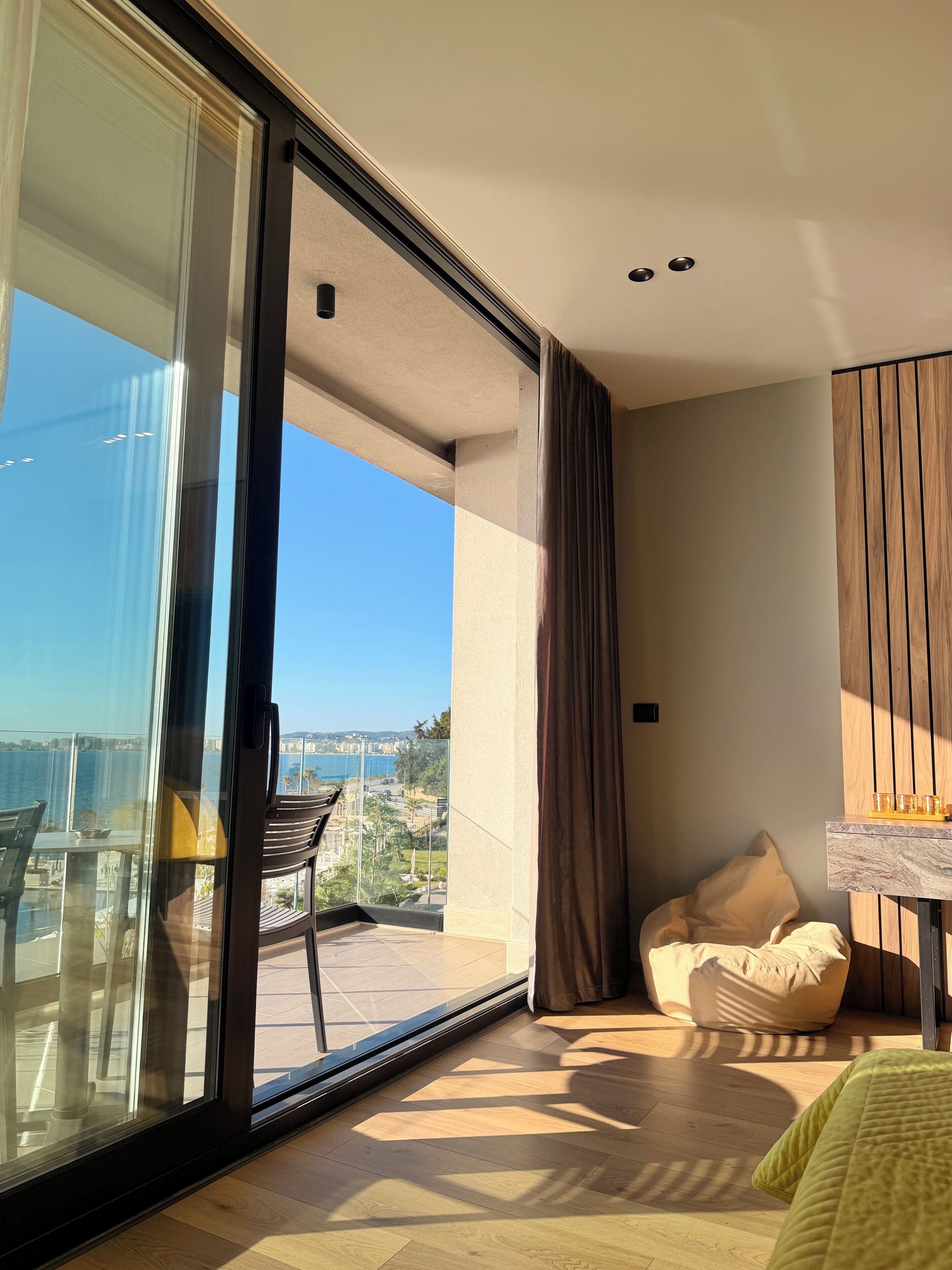 A modern room with a large sliding glass door leading to a balcony with a view of the sea and distant buildings.