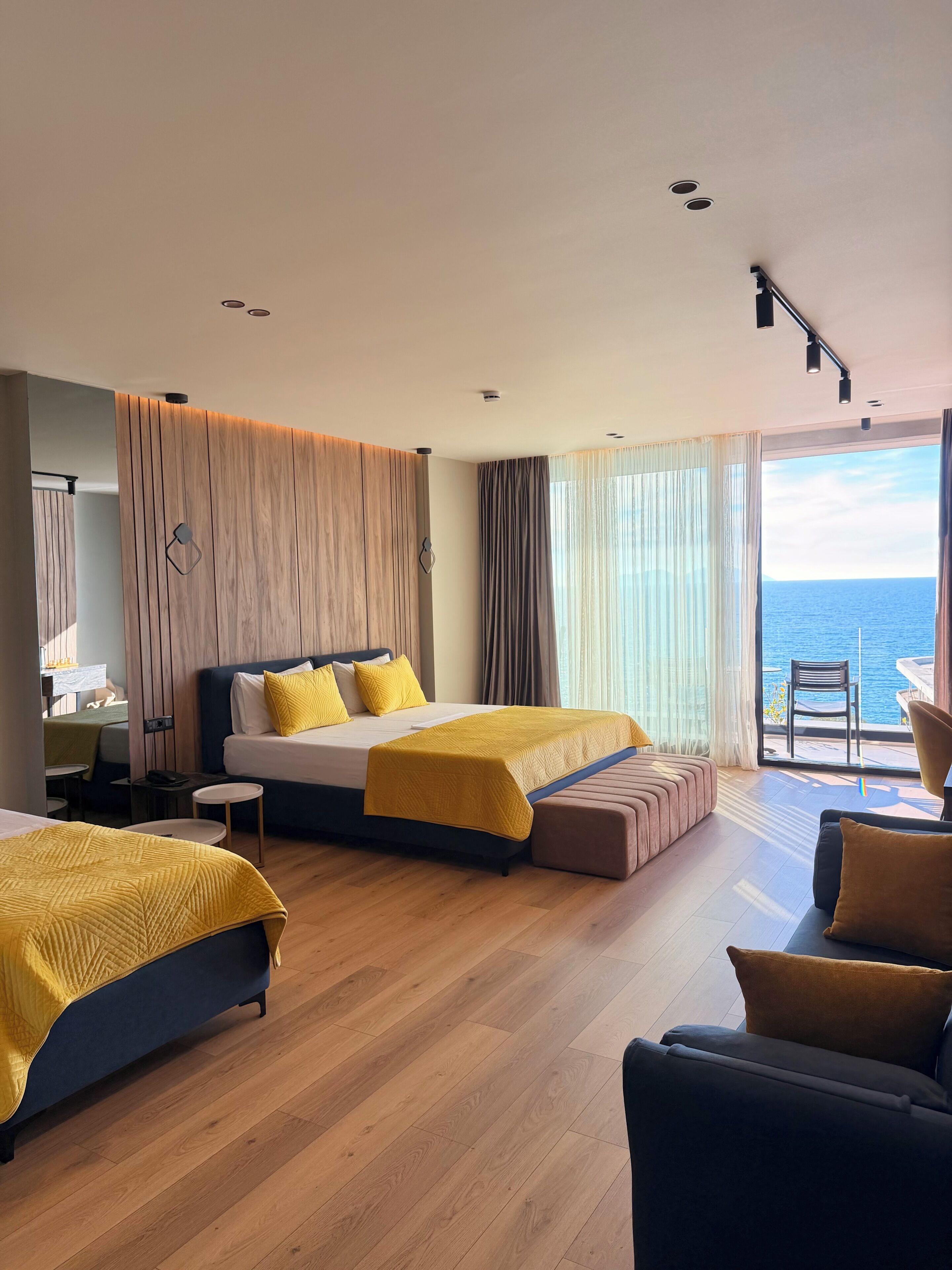 A hotel room with two beds, a sofa, a dining area, and a balcony with a view of the sea.