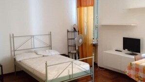 Un dormitorio con estructura de cama metálica, una mesita de noche pequeña, un televisor sobre un mueble bajo y una ventana con cortinas.